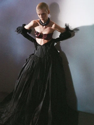 Model with short blonde hair in a voluminous black gown, sequined bralette, and long black gloves against a shadowed wall.