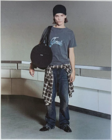 Model wearing a beanie, faded graphic t-shirt, plaid shirt tied at the waist, and dark jeans while holding a circular black bag.