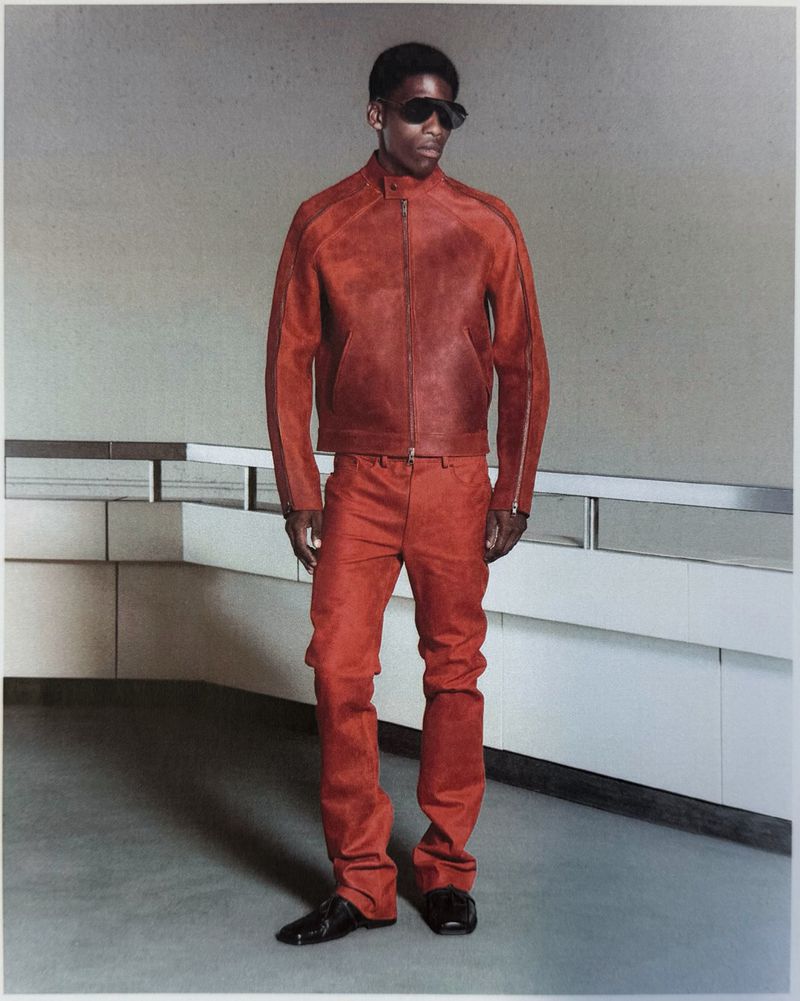 Model in a rust-red leather jacket, matching trousers, and dark sunglasses standing in a minimalist interior.