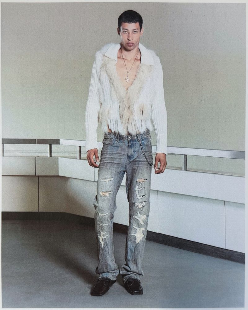 Model wearing a white faux fur-trimmed knit jacket and distressed denim jeans standing against an interior railing.