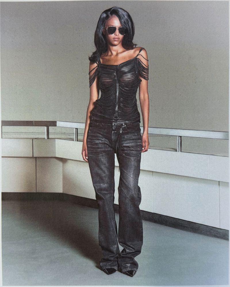 Model wearing dark sunglasses, black fringe top, and dark jeans posing in an interior space with a railing