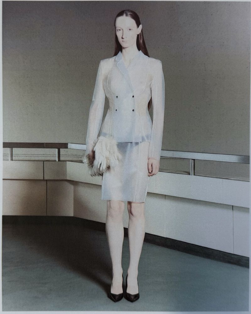 Model wearing a sheer translucent blazer and skirt suit with black pointed pumps in an architectural hallway.