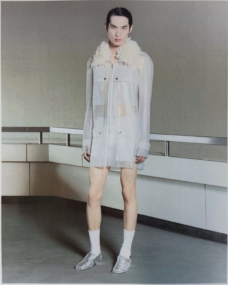 Model wearing a sheer zip-up coat with a textured fur collar, white socks, and silver metallic loafers standing against a railing.