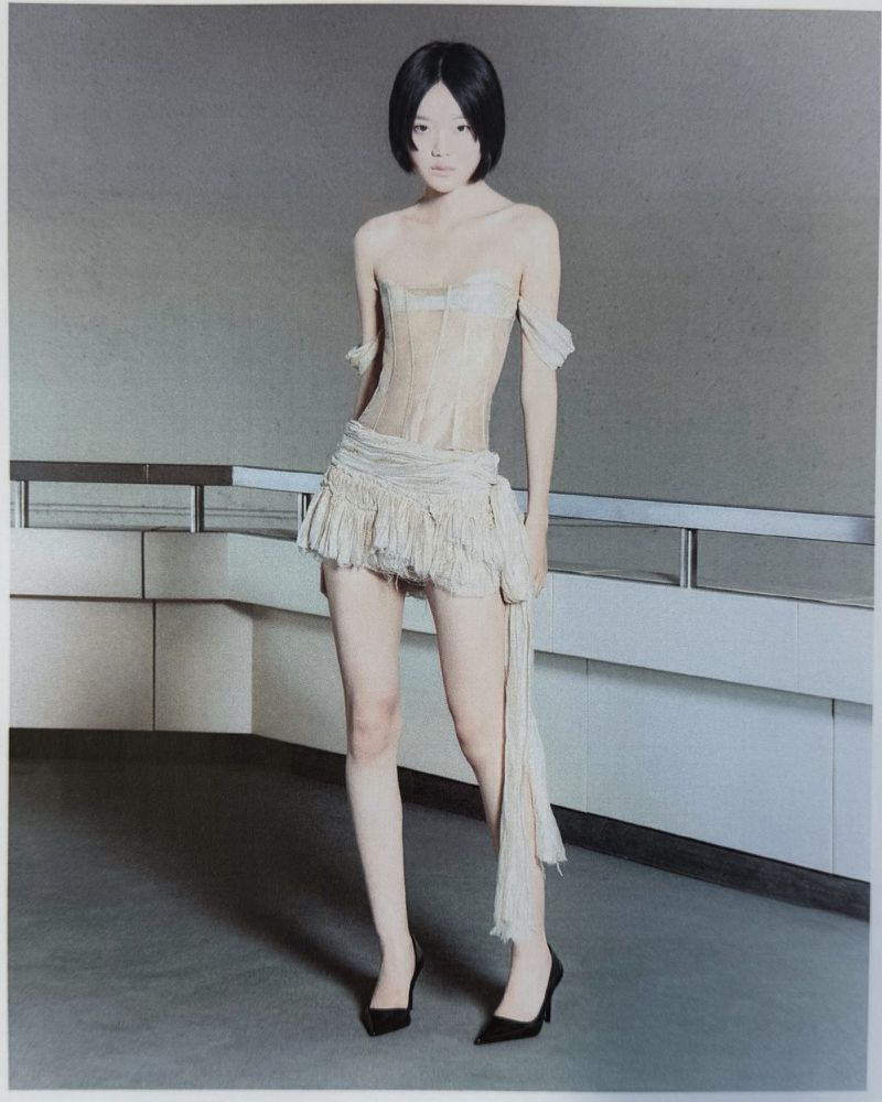 Model with a black bob wearing a sheer beige corset top, fringe mini skirt, and black pointed heels standing by a railing.