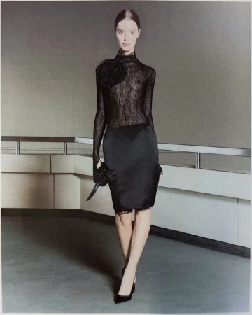 Fashion model in a sheer black lace long-sleeve top and black satin pencil skirt standing before an indoor architectural railing.