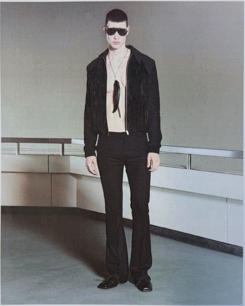 Model wearing an open black jacket, black flared trousers, sunglasses, and a pendant necklace for the Unvain S/S 27 Lookbook.