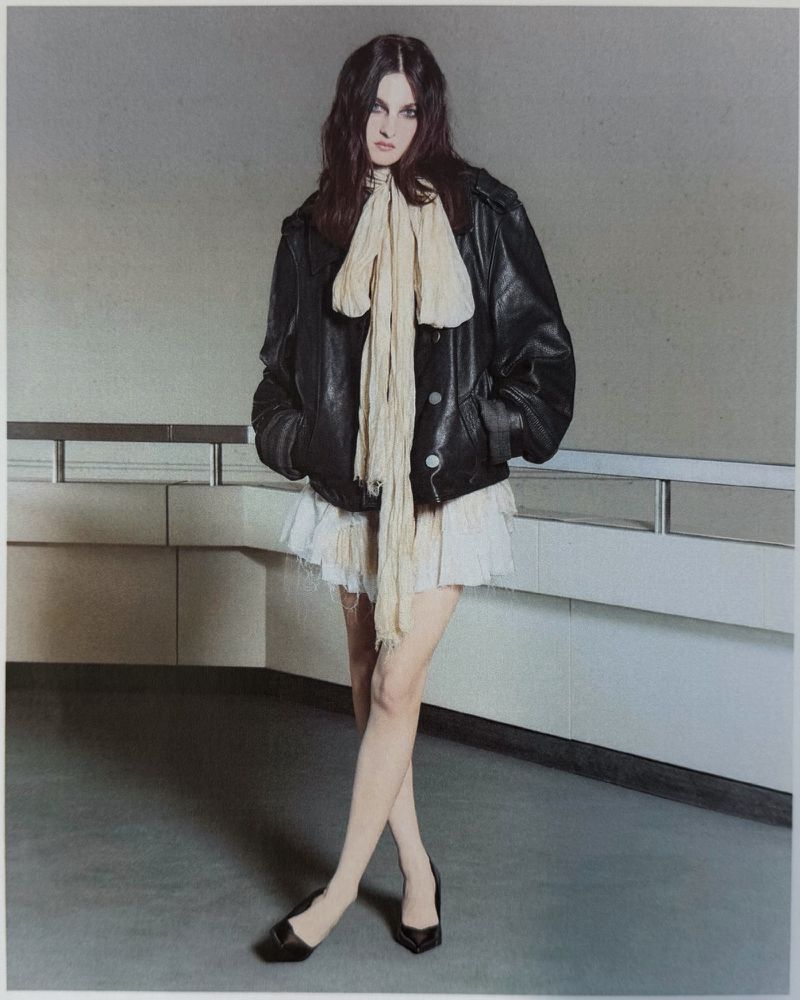 Model posing in an oversized black leather jacket with a long cream scarf and ruffled mini skirt by an architectural railing.