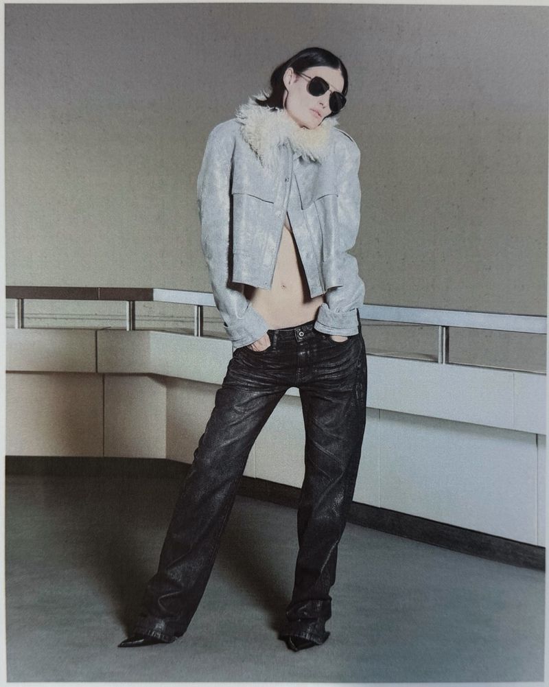 Model wearing dark sunglasses, a cropped shearling-collar jacket, and dark denim jeans posing beside an interior railing.