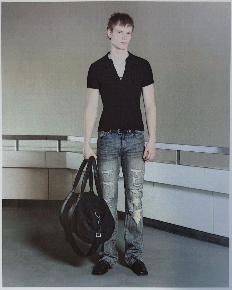 Young man standing with a black duffle bag, wearing a black short-sleeve polo and distressed faded jeans in front of a metal railing.