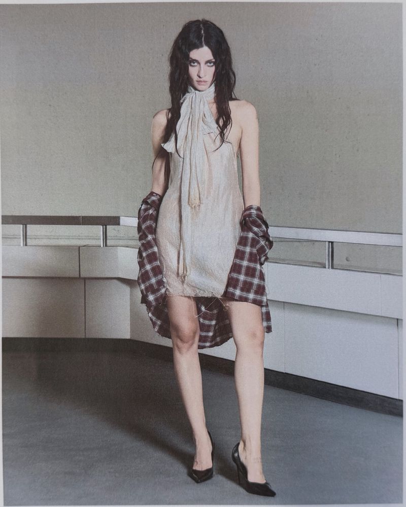 Model wearing a sheer halter dress with neck scarf, draped plaid shirt, and black heels standing by a concrete railing.