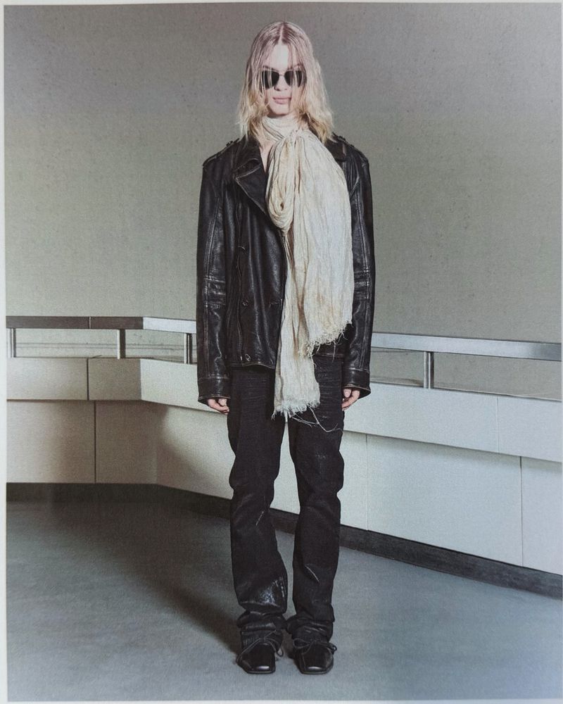Model wearing dark sunglasses, a black leather jacket, a cream fringed scarf, and dark trousers standing in a minimalist corridor.