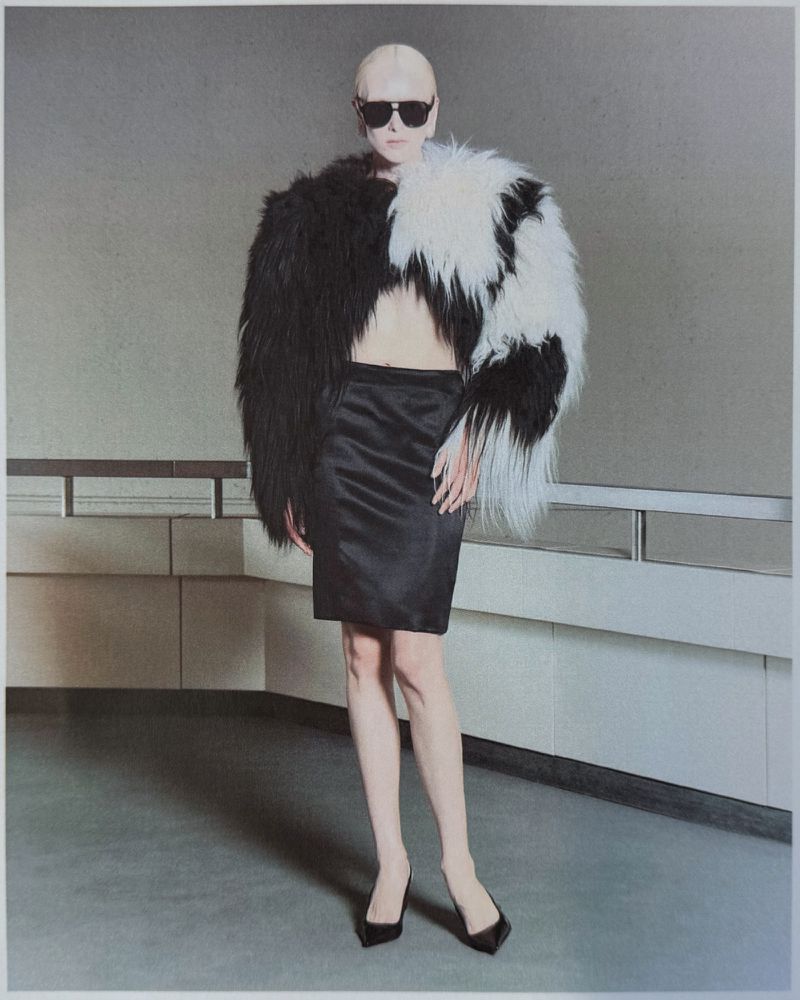 Model wearing black sunglasses, a two-tone black and white shaggy fur cape, a black satin pencil skirt, and pointed black pumps.