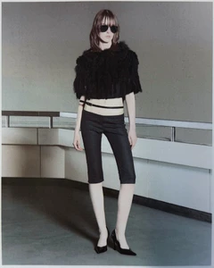 Model wearing dark sunglasses, a black textured crop top, black knee-length shorts, and heels against a concrete wall.
