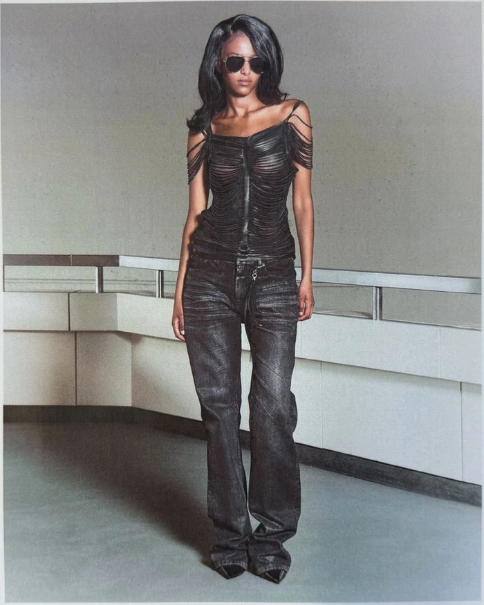 Model wearing dark sunglasses, black fringe top, and dark jeans posing in an interior space with a railing