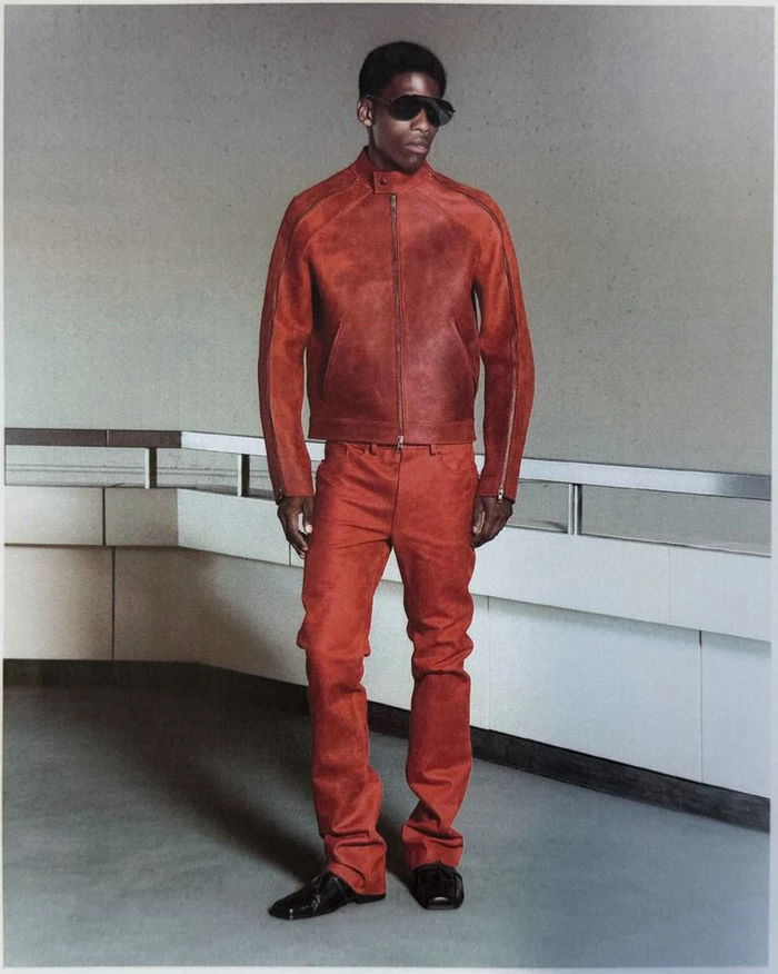 Model in a rust-red leather jacket, matching trousers, and dark sunglasses standing in a minimalist interior.