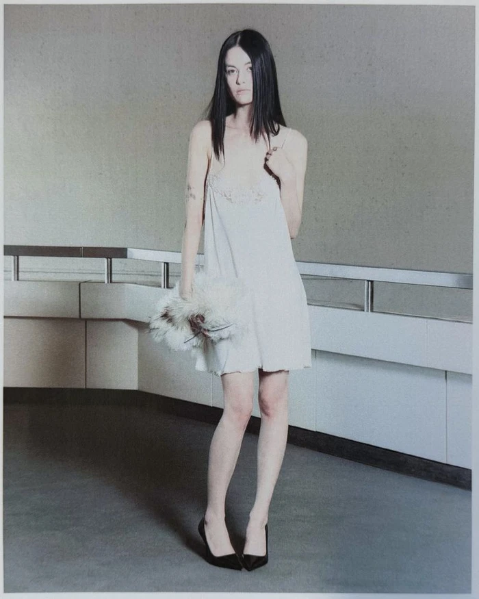 Model with straight dark hair wearing a white mini slip dress and black pointed pumps while holding a white feathered clutch.