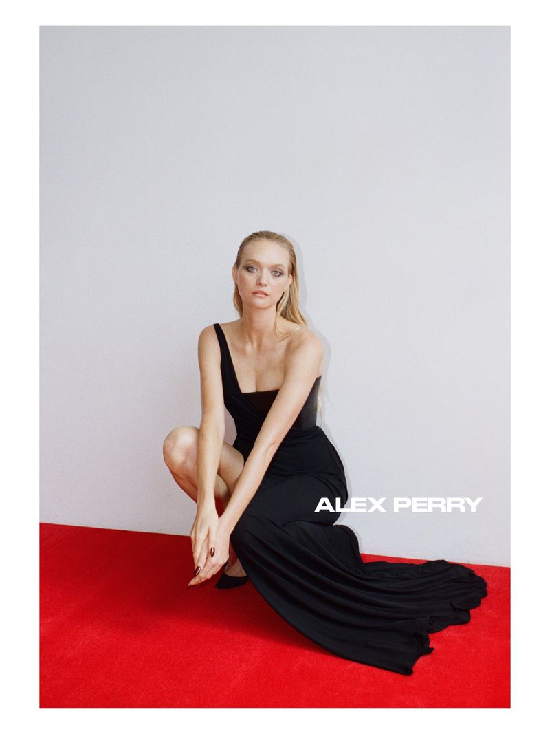 Model Gemma Ward wearing a long black evening gown crouching on a red carpet against a light grey wall for Alex Perry