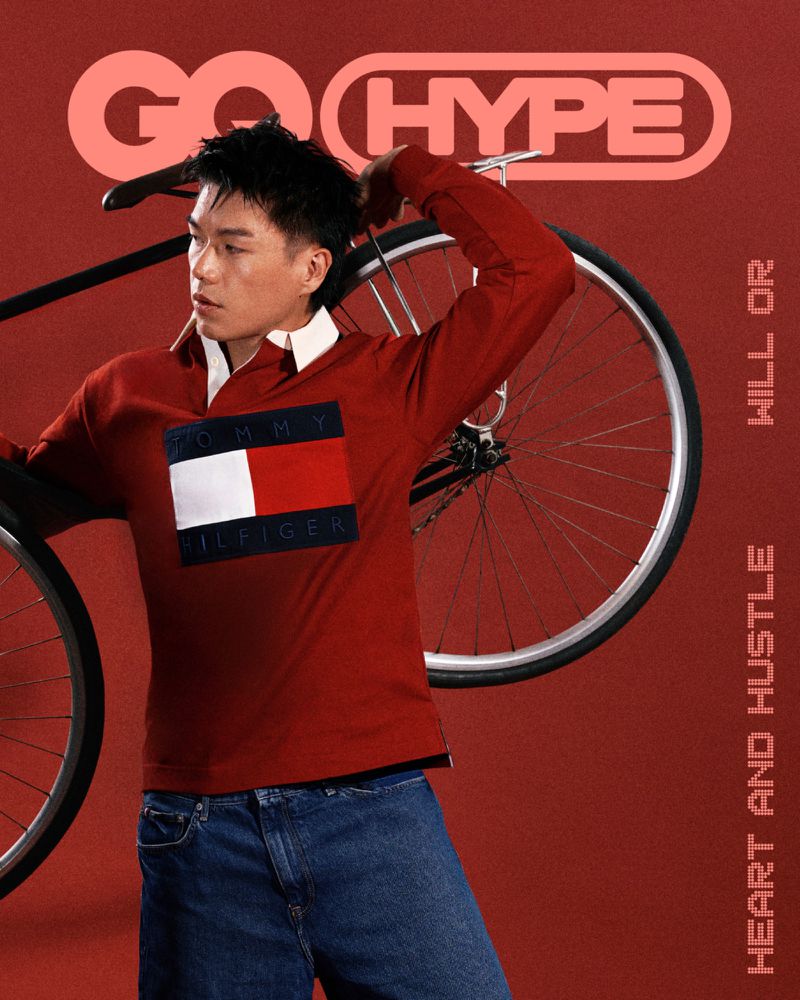 Man holding a bicycle behind his back wearing a red long-sleeve Tommy Hilfiger polo shirt against a red backdrop for GQ Hype.
