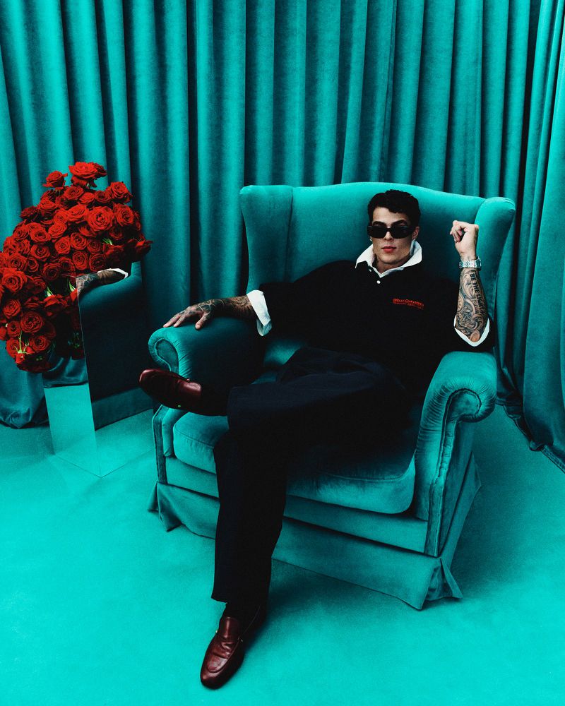 Man in armchair, sunglasses, and roses in a turquoise studio setting. Tattoos, dark polo shirt, and leather shoes in a staged studio shot.