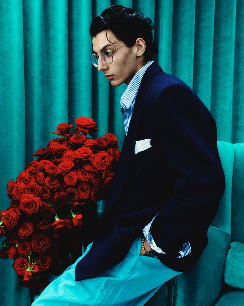 Portrait of a young adult man wearing glasses, a blazer, and a striped shirt, posing with a large bouquet of red roses.