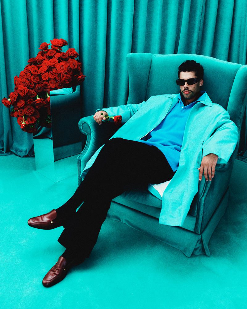 Adult male in sunglasses and coat reclines in turquoise wingback chair with red roses in a matching teal studio setting.