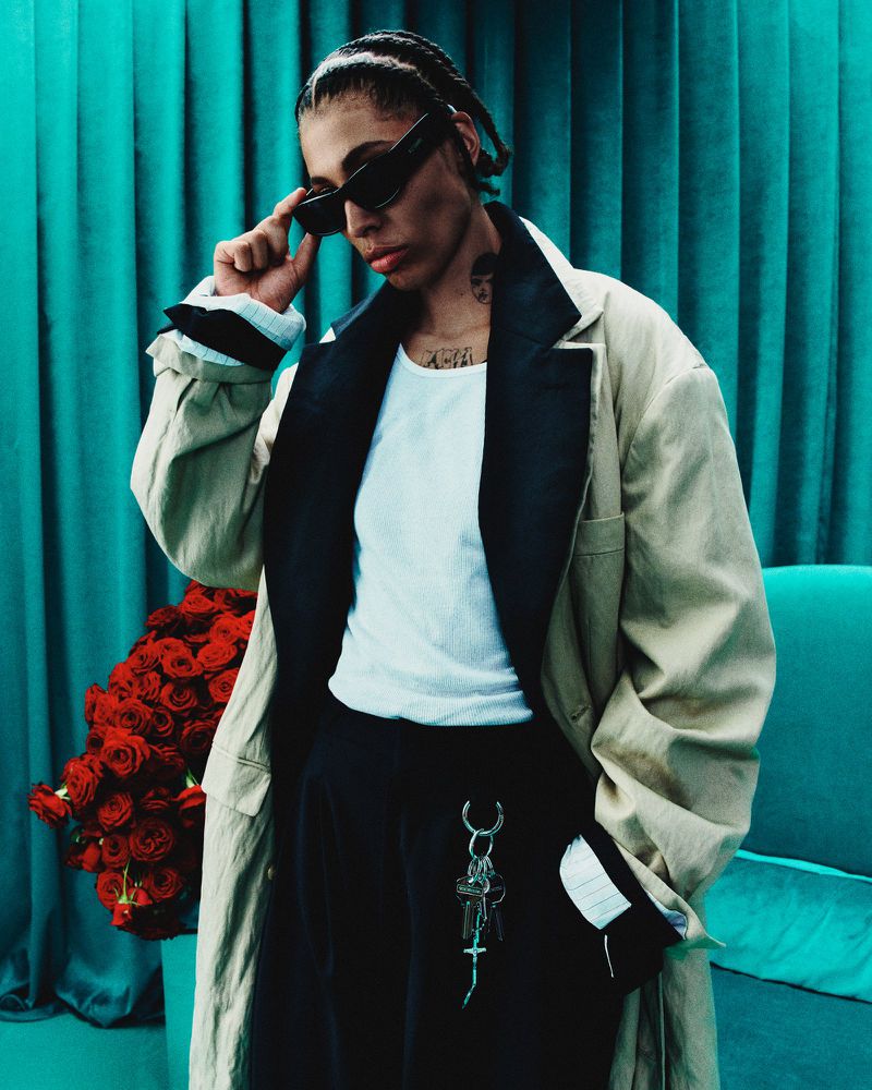 Stylish woman in sunglasses and trench coat posing with roses against turquoise curtain, capturing a blend of fashion and elegance.