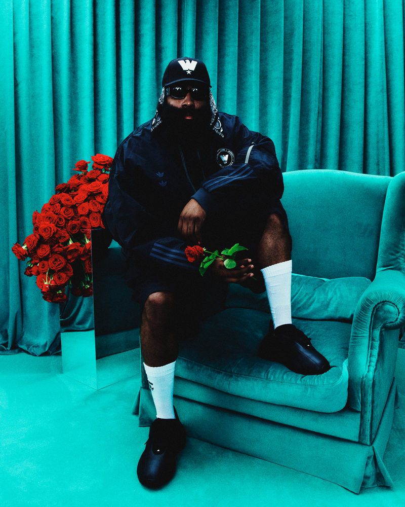 Bearded man in a black tracksuit, hat, and white socks sits on a teal couch, holding a red rose against a matching teal background.