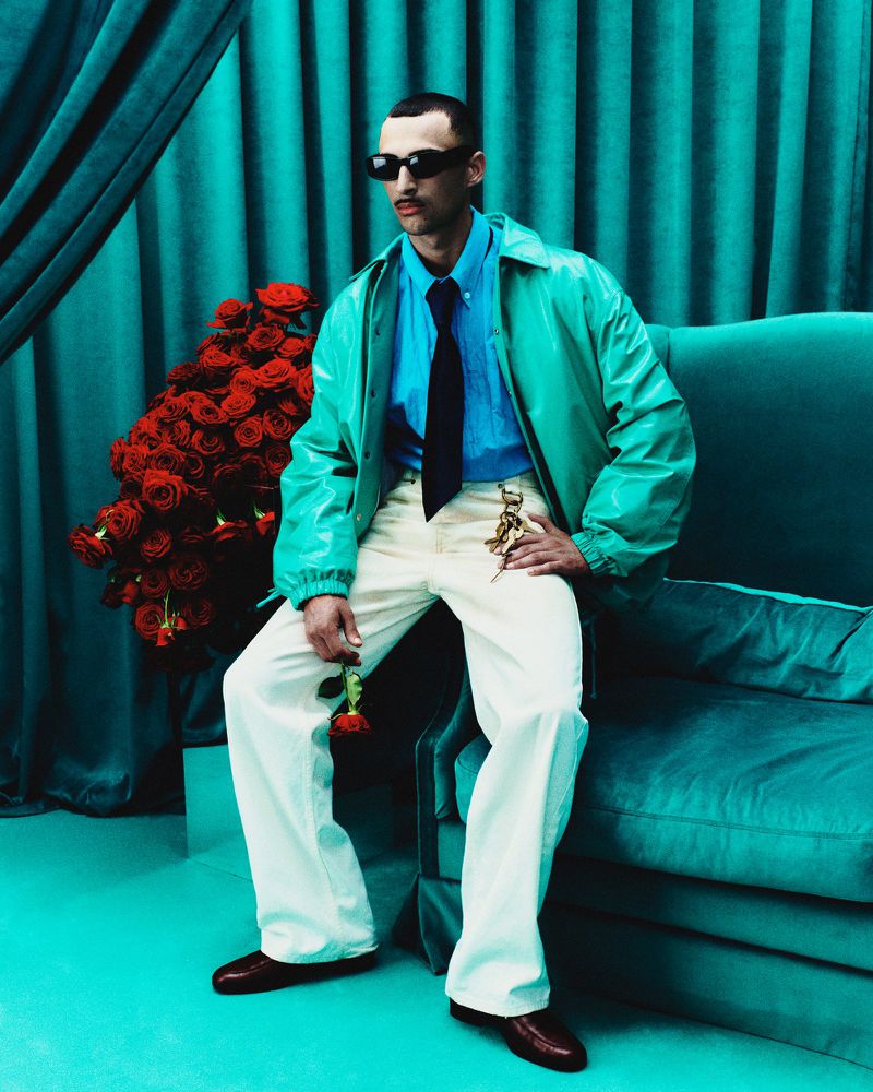 Adult male in light green jacket poses on green velvet couch next to red roses against a green curtain in studio setting.