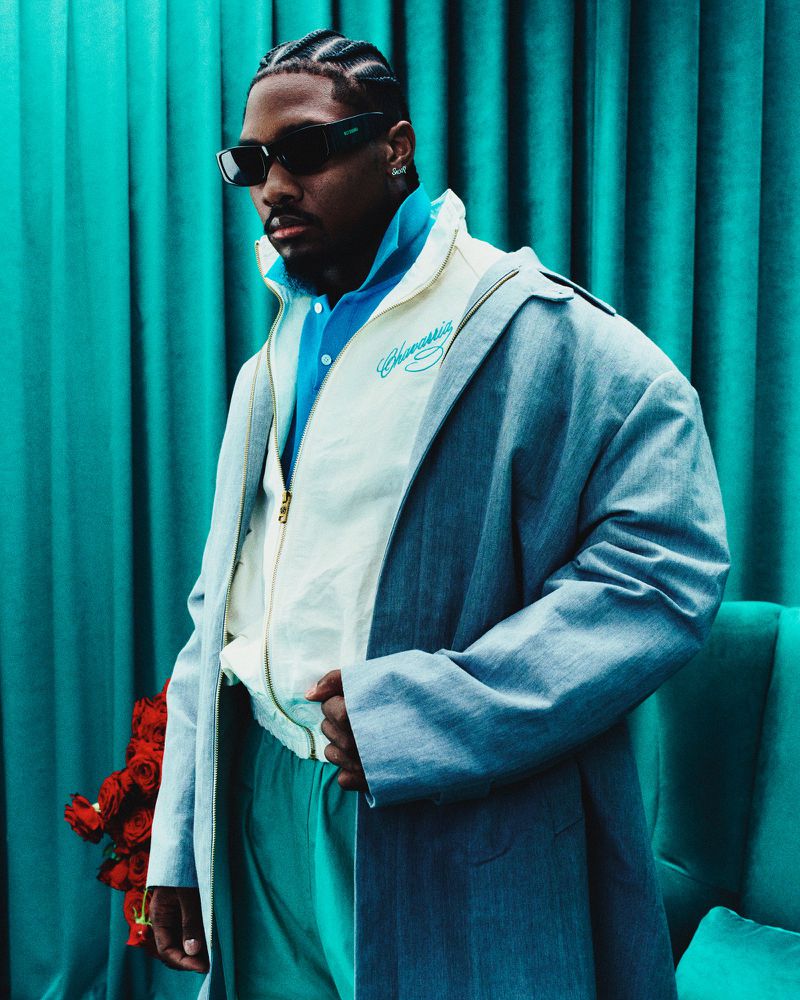 Portrait of an adult male wearing sunglasses and a jacket, posing in front of a turquoise curtain with a bouquet of red roses.