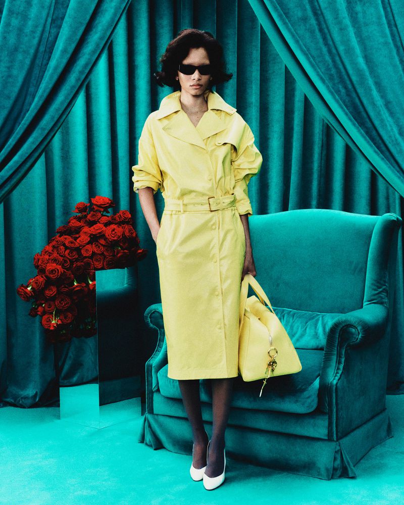 A woman models a yellow trench coat with a matching handbag in front of a turquoise curtain and a vase of red roses.