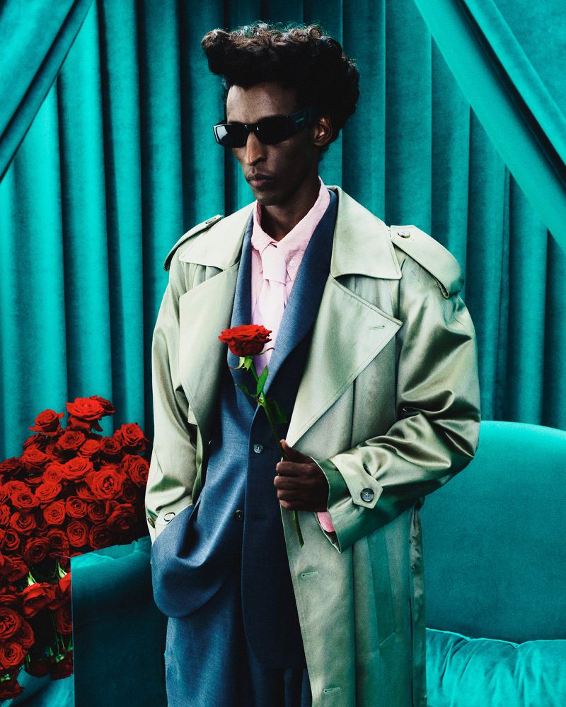 Man in suit and trench coat holds a red rose, posing against a turquoise draped background with roses, fashion forward, formal attire.