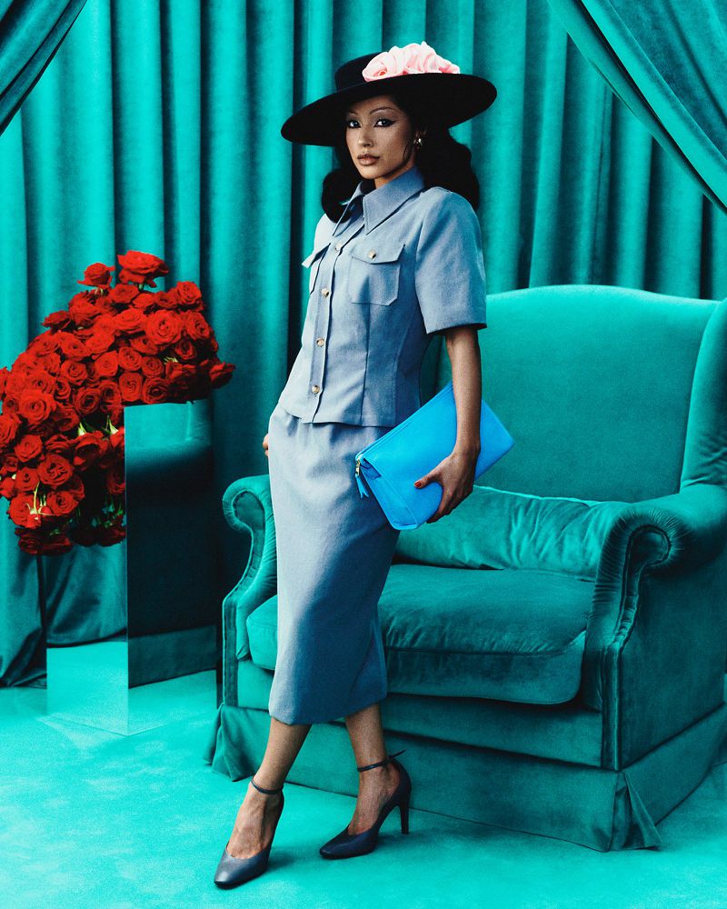 Elegant woman in a light blue suit and wide-brimmed hat poses with a blue clutch against a teal backdrop, complemented by vibrant red roses.