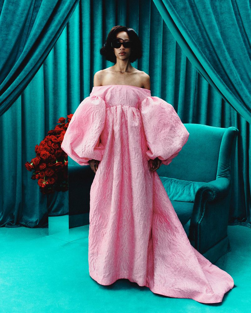A woman with sunglasses wears a pink gown in a studio with a turquoise backdrop and an armchair next to roses, fashion concept.