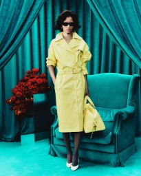 Model wearing a yellow belted trench coat, sunglasses, and white pumps standing before a teal velvet armchair and drapery.