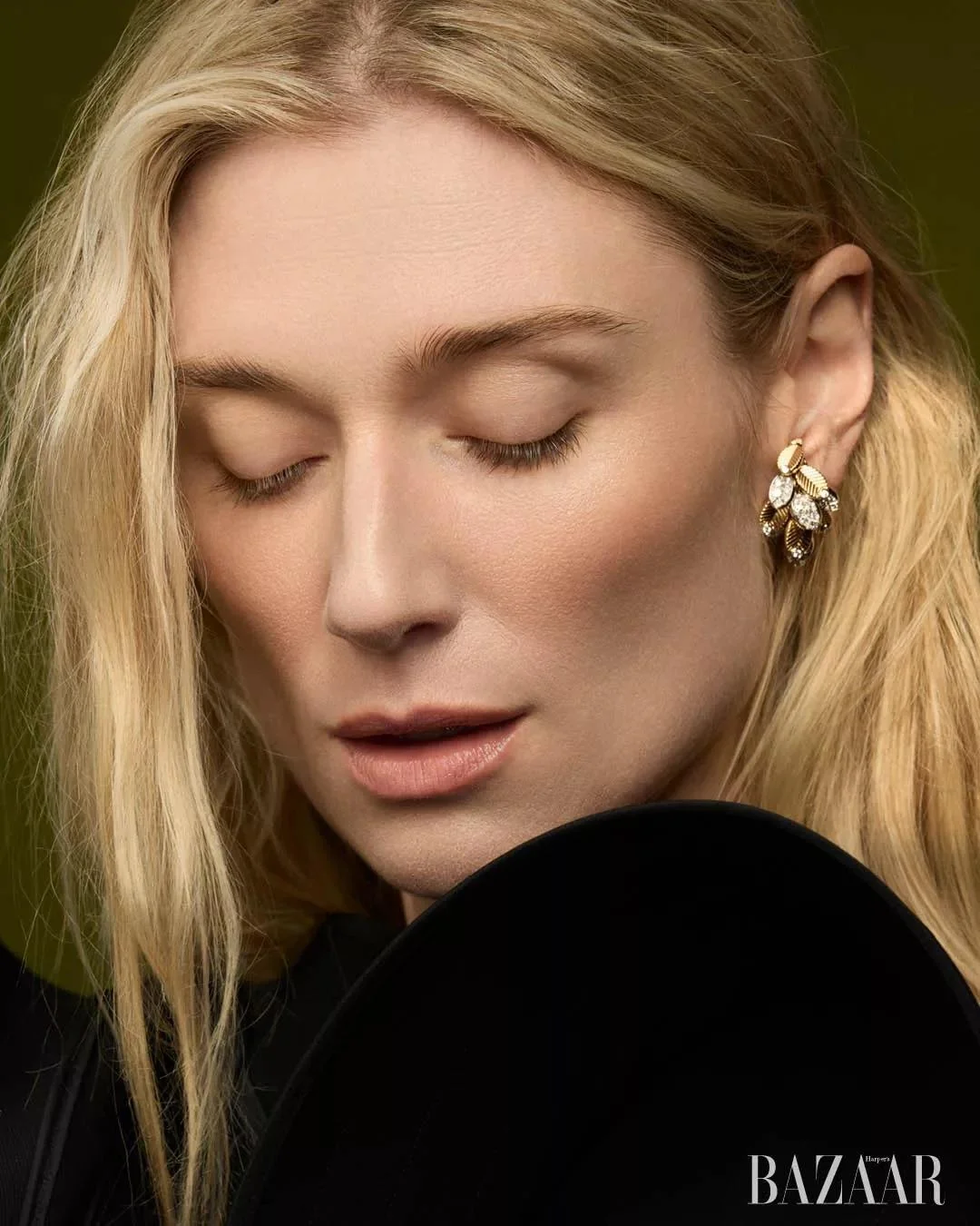 Elizabeth Debicki with closed eyes wearing an ornate gold leaf earring and black top against an olive background.