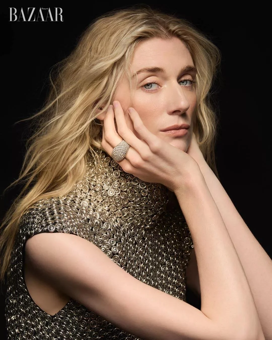 Portrait of actress Elizabeth Debicki wearing a metallic textured top and diamond ring against a plain black background.