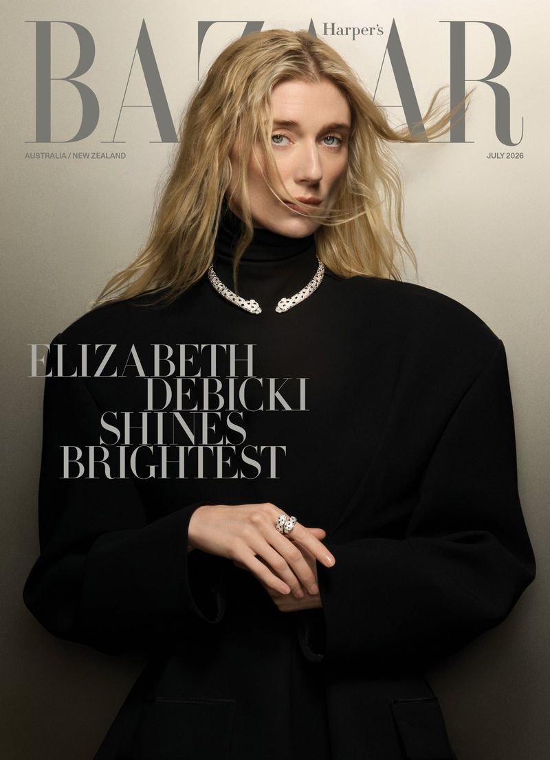 Elizabeth Debicki poses on a magazine cover wearing a structured black turtleneck coat and sculptural diamond jewelry against a grey studio backdrop.