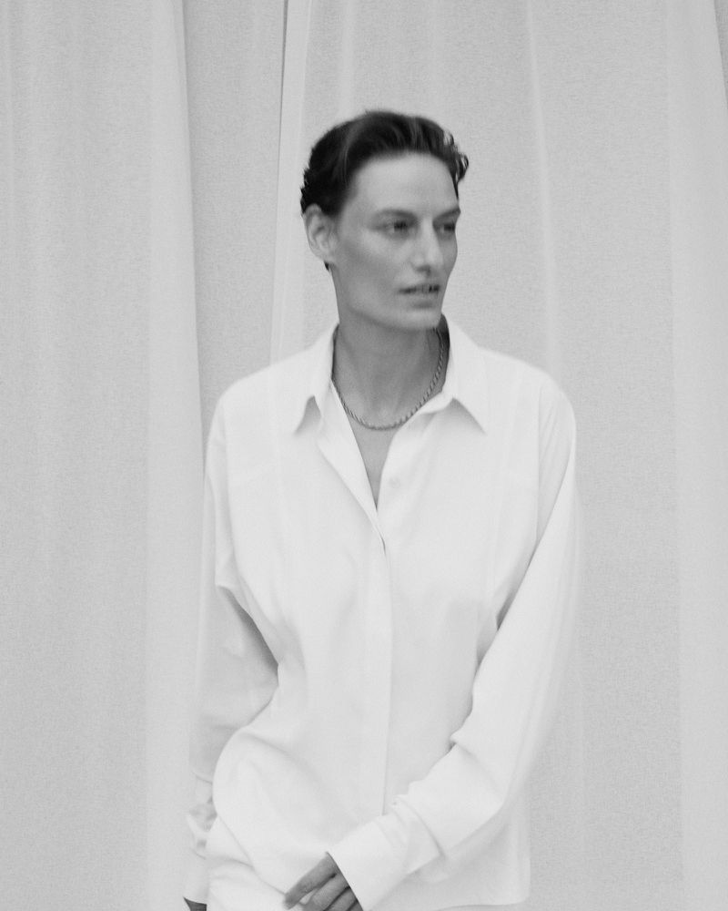 Veronika Kunz posing in an oversized white button-down collared shirt with draped fabric in the background.