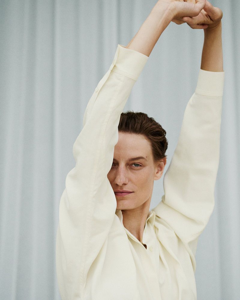 Veronika Kunz raising her arms while wearing a cream long-sleeve button-up shirt against a pleated curtain backdrop.