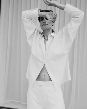 Model Veronika Kunz posing in an unbuttoned white shirt and matching trousers against pleated drapery.
