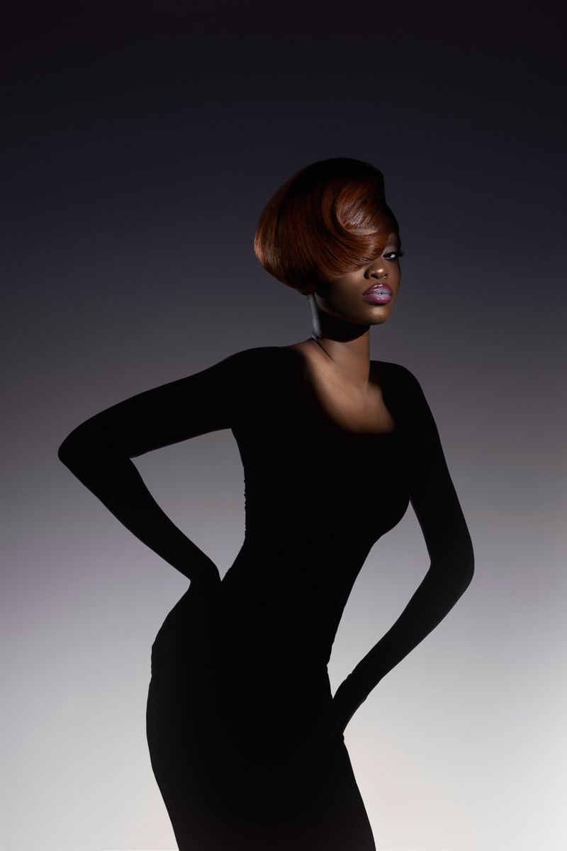 Elegant studio portrait of a Black woman with a sleek auburn hairstyle, wearing a fitted black dress, hands on hips, against a gradient gray backdrop.