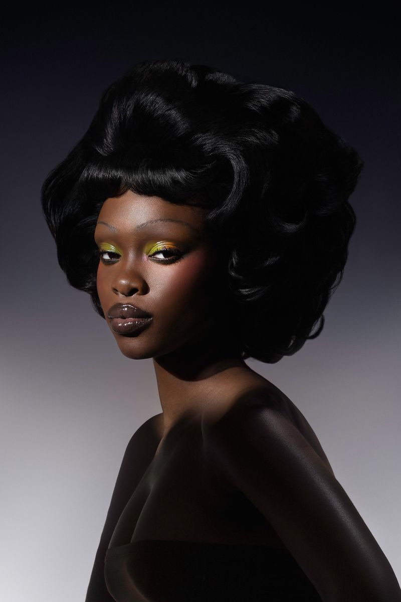 Close-up portrait of an African American adult female with dramatic makeup, glossy lips, and voluminous black hair against a grey background.