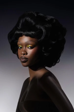 Close-up portrait of an African American adult female with dramatic makeup, glossy lips, and voluminous black hair against a grey background.