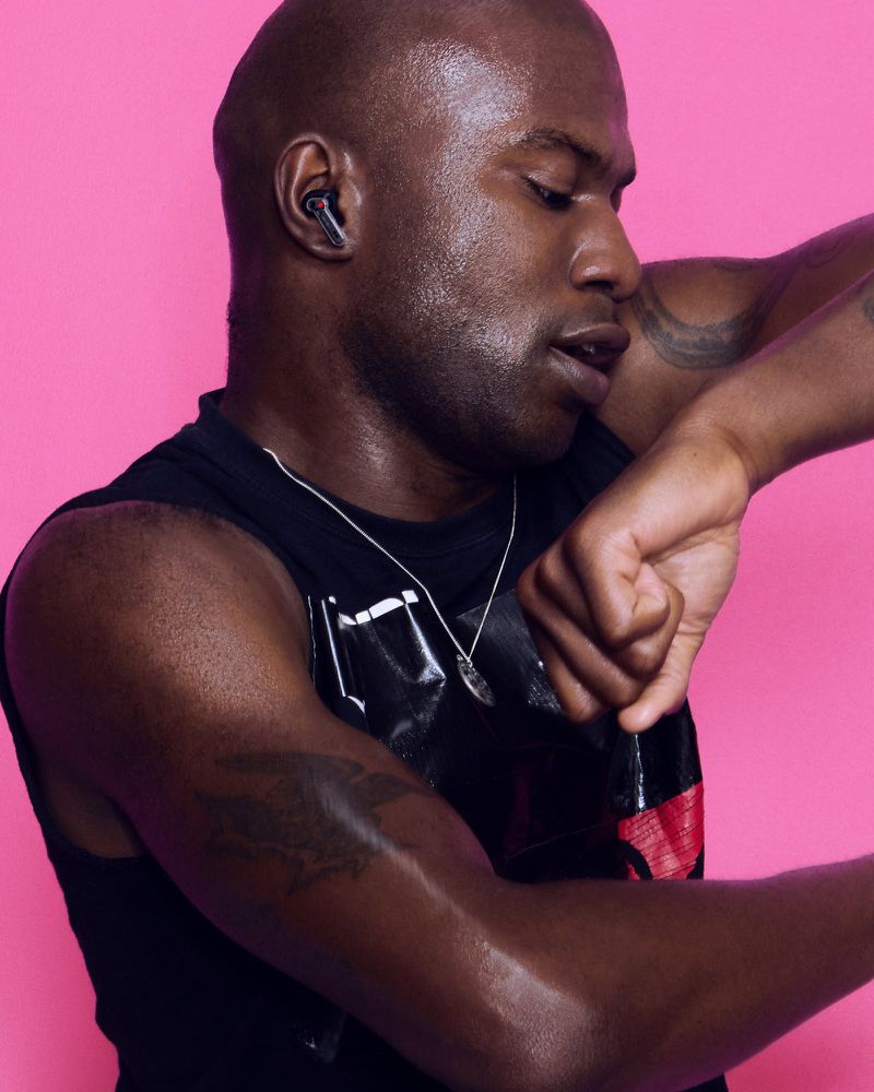 Black man with glistening skin wearing a transparent wireless earbud and black sleeveless top against a bright pink background.