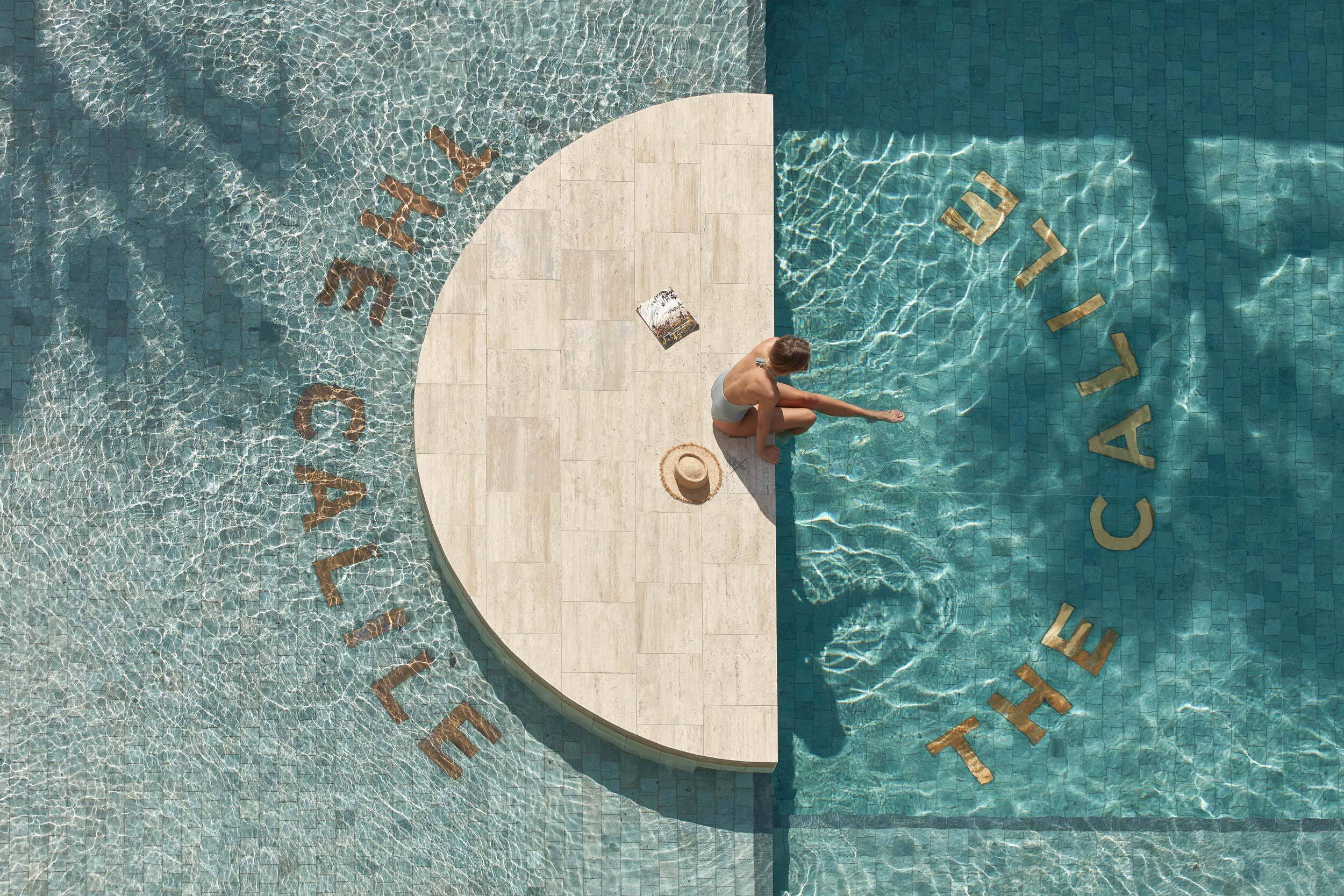 Overhead view of a woman sitting on a semicircular stone ledge in a turquoise swimming pool beside a sunhat and magazine.
