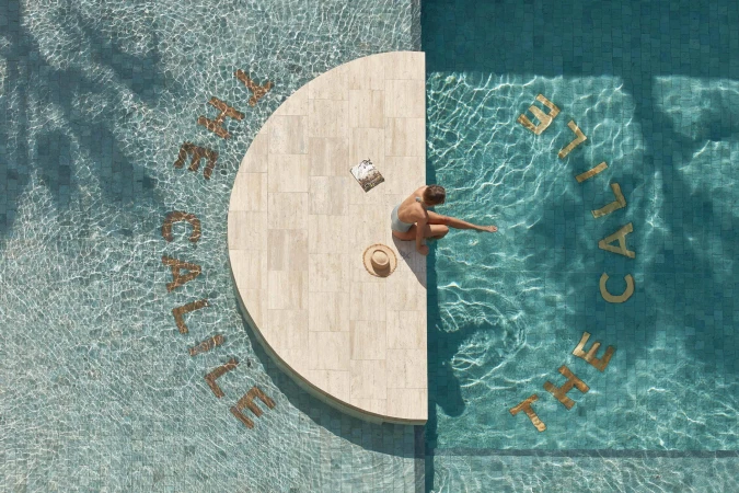 Overhead view of a woman sitting on a semicircular stone ledge in a turquoise swimming pool beside a sunhat and magazine.