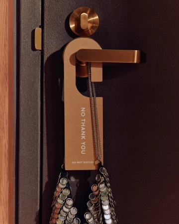 A tan door hanger reading No Thank You on a brass handle alongside a sequin purse against a dark door.
