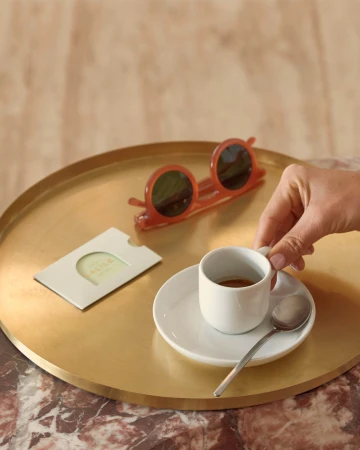 A hand holds a white espresso cup on a circular brass tray alongside round amber sunglasses and a hotel keycard holder.
