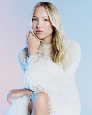 Model Lila Moss resting her hand on her chin wearing a sheer white long-sleeve dress with feather trim against a pastel gradient.