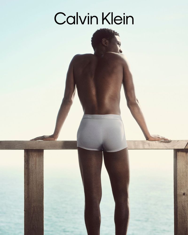 Shirtless black male model seen from behind leaning on a wooden railing against a clear sky for Calvin Klein